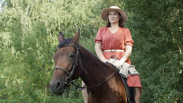 Beautiful horsewoman in dress and hat sitting horseback while forest walk. Happy woman sitting astride brown horse on summer forest trees background