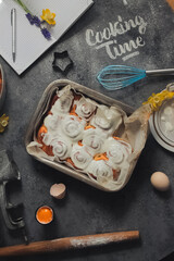 Homemade cinnamon rolls buns with white cream icing ,word cooking time made of white flour, blue whisk and egg shell on grey dark concrete background, bake paper, plate, notebook and pen, daffodil