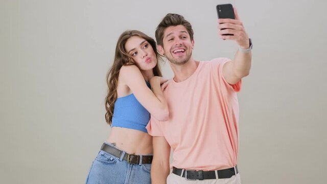 A pleased positive young couple is taking selfie photo on the smartphone standing isolated over gray background in studio