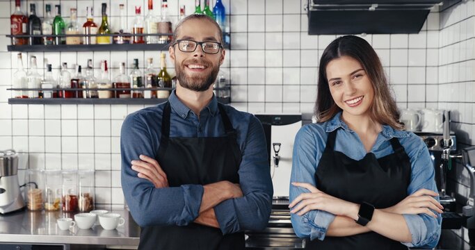 Portrait young Caucasian cheerful couple of bartenders in aprons looking at each other and smiling to camera happily. Happy waiter and waitress posing in cafe. Joyful man and woman, barristas at work.