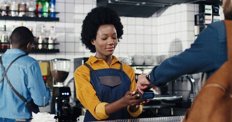 Multiethnic bartender woman and client man. Caucasian male paying with paypass, credit card. Mixed-races. Passing payment at counter. Pay for drink or food in cafe. African American waitress.