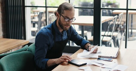 Caucasian young man sitting at table and doing business calculations. Man in glasses calculating...