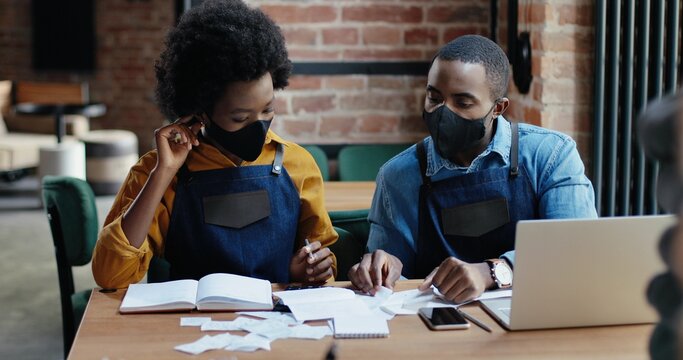 African American couple in masks doing business calculations of restaurant and checking bills. Calculating spends and damages. Reopen after lockdown. Counting at laptop computer Small entrepreneurship