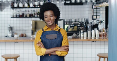 Portrait of African American young happy beautiful woman barrista in apron standing at counter in bar, smiling to camera and crossing hands. Waitress posing in cafe with drinks equipment on background