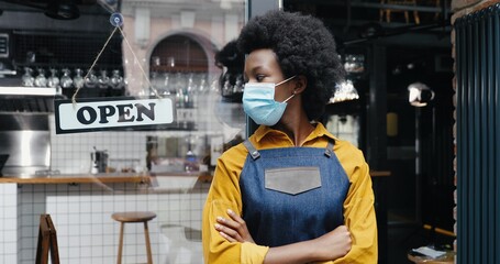 Portrait of beautiful African American young woman waitress in apron and medical looking to camera with table open at cafe entrance outdoor. Pretty barrista standing at bar door with board open Reopen