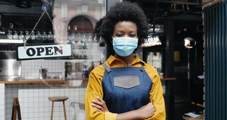 Portrait of beautiful African American young woman waitress in apron and medical looking to camera with table open at cafe entrance outdoor. Pretty barrista standing at bar door with board open Reopen