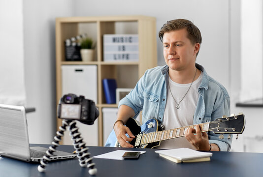 video blogging, music and people concept - young man or musician with camera videoblogging and playing guitar sitting at table at home