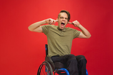 Closing ears. Caucasian young disabled man's portrait on red studio background. Beautiful model in casual style. Concept of human emotions, facial expression, sales, ad, inclusion. Copyspace.