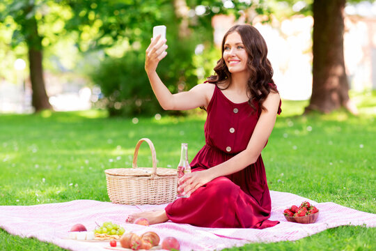 leisure and people concept - happy smiling woman with smartphone and fizzy drink in bottle sitting on picnic blanket and taking selfie at summer park