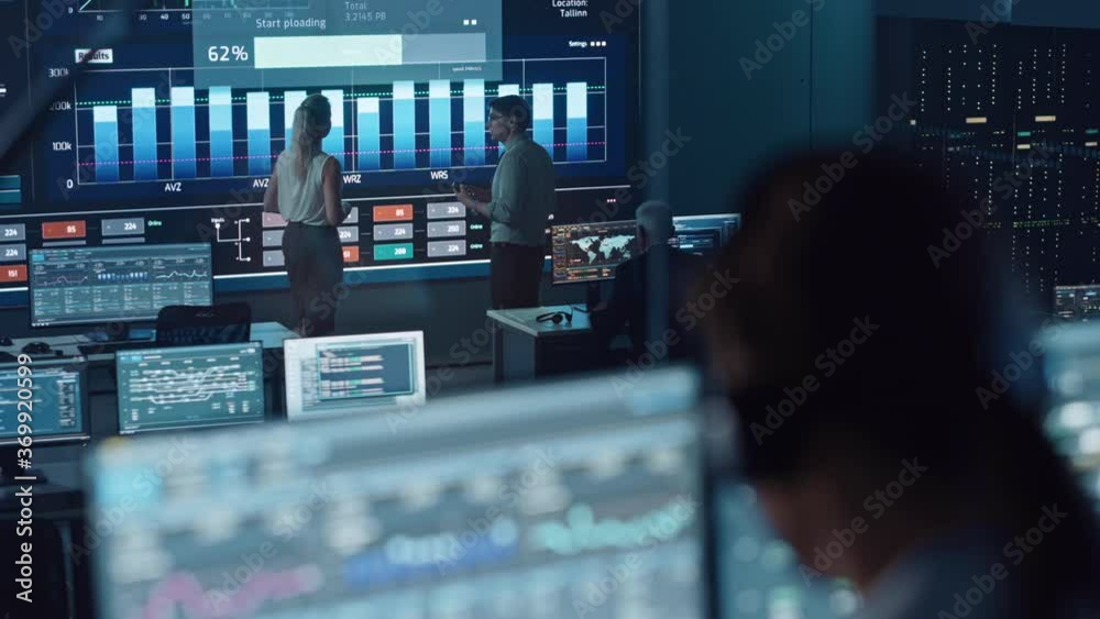 Poster Two Software Engineers Talking Next to a Big Screen in a Modern Monitoring Office with Live Analysis Feed with Charts. Monitoring Room Big Data Scientists and Managers Sit in Front of Computers.