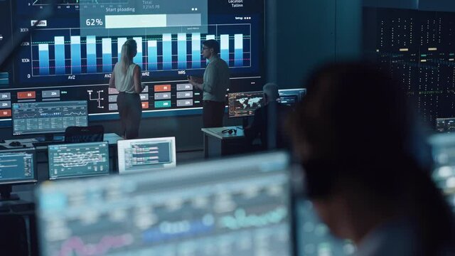 Two Software Engineers Talking Next to a Big Screen in a Modern Monitoring Office with Live Analysis Feed with Charts. Monitoring Room Big Data Scientists and Managers Sit in Front of Computers.