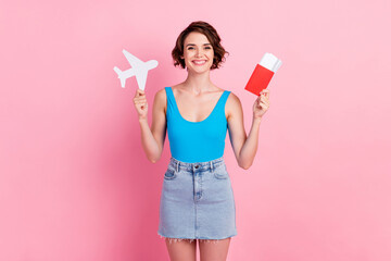 Photo of cute lovely young tourist lady hold paper plane tickets passport cheerful smile packing suitcases prepare journey wear denim mini skirt blue singlet isolated pink color background