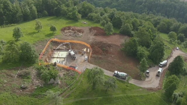 AERIAL: Cement truck drives up the road while another vehicle pours the concrete foundation of a house in the idyllic green countryside in Slovenia. Cement pouring in progress at a construction site.