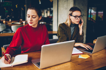 Two skilled graphic designers working remotely at modern netbooks connected to wireless internet in coworking.Blonde student calling on smartphone while colleague noting text in notepad from website