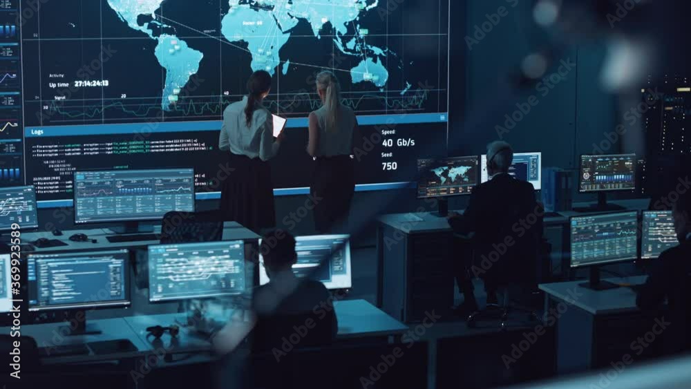 Poster Two Software Engineers Talking Next to a Big Screen in a Modern Monitoring Office with Live Analysis Feed with Charts. Monitoring Room Big Data Scientists and Managers Sit in Front of Computers.