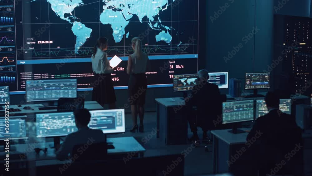 Poster Two Software Engineers Talking Next to a Big Screen in a Modern Monitoring Office with Live Analysis Feed with Charts. Monitoring Room Big Data Scientists and Managers Sit in Front of Computers.