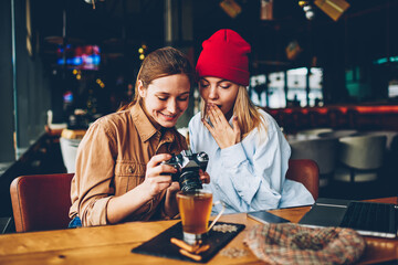 Wondered stylish teenager with cheerful friend female photographer dressed in casual outfit watching shocked photos on vintage camera during meeting in cafe interior enjoying recreation time