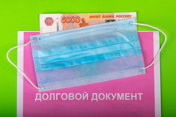 Protective medical mask lies on debt reporting document with the inscription in Russian "debt document" and russian money on a green background. 