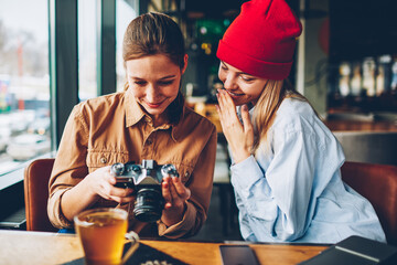 Cheerful female students watching made positive photos on display of vintage camera resting together in stylish cafeteria.Amazed hipster teenagers making editing of funny pictures during free time