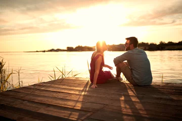 Fotobehang young couple sitting on wooden platform by the lake, looking each other, talking at sunset © luckybusiness