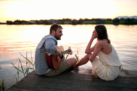 caucasian female enjoying in company of a beardy male playing guitar by the lake at sunset