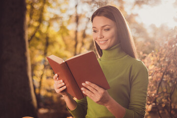 Photo of pretty charming intelligent lady come autumn park read science fiction novel sit among trees comfortable lonely place dreamy beaming smiling wear green turtleneck outdoors