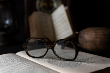 Old worn Glasses resting on an opened book with books and clock in background
