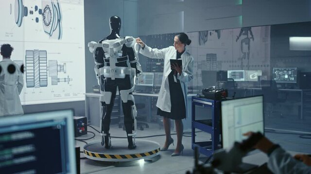 In Robotics Development Laboratory: Female Scientist Uses Digital Tablet Computer, Work on a Robotics Exoskeleton Prototype. Engineers Designing Exosuit to Help Disabled People, Workers Lift Goods 