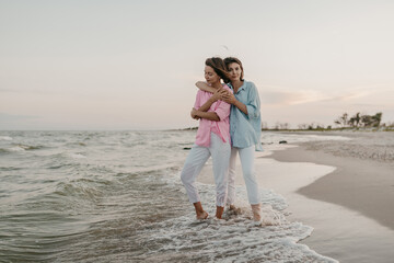 pretty two young women friends having fun on the sunset beach, queer non-binary gender identity, gay lesbian love romance, boho summer vacation style wearing jeans