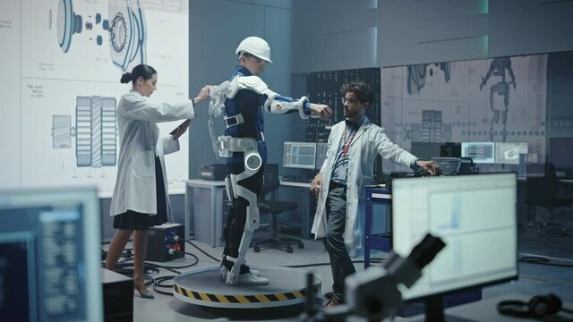 In Robotics Development Laboratory: Engineers and Scientists Work on a Bionics Exoskeleton Prototype with Person Testing it. Designing Wearable Exosuit to Help Disabled People, Warehouse Workers
