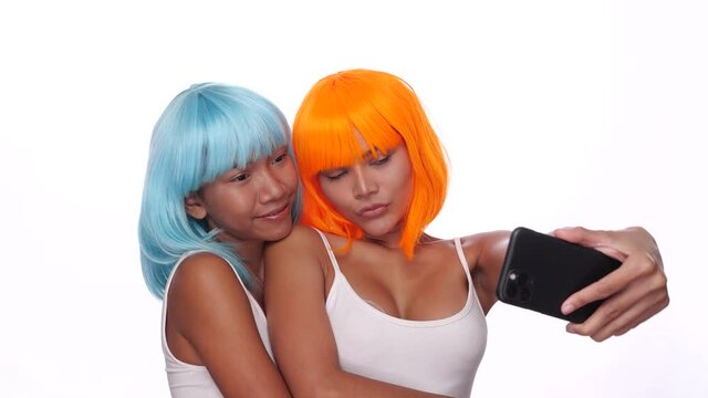 Portrait of two beautiful happy Asian women friends in bright blue and orange wig doing selfie photo with smartphone over white background