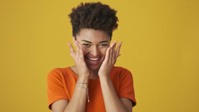 Cheerful attractive african woman hiding behind her hands then open her face and having fun over yellow background