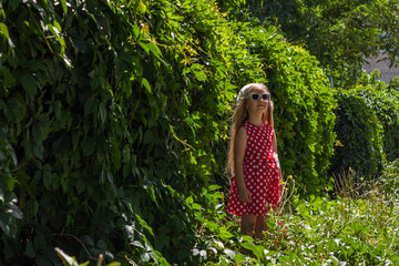 Beautiful happy girl in a red polka dot dress smiling on wild grape background. Cute joyful child model with long blonde hair in sunglasses and white hat on natural green leaves backdrop outdoor.