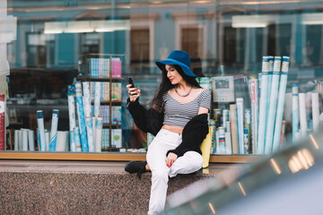 Young glamorous woman connects to the Internet via smart phone while sitting near window of bookstore,funky female watching video on cell telephone while relaxing after walking during recreation time