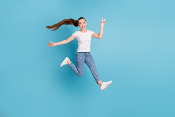 Full size photo of energetic excited crazy girl teen enjoy free time jump make v-sign wear white t-shirt gumshoes isolated over blue color background