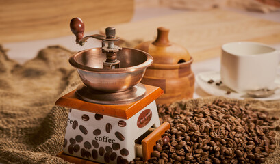 manual coffee grinder with scattered coffee beans, blur background sugar bowl and a cup with a saucer