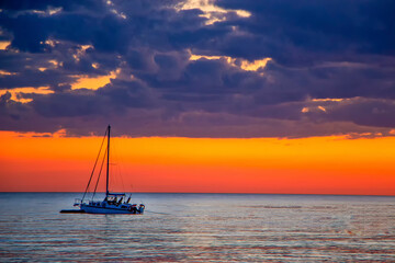 Yacht in the sea at sunset, against a beautiful sky with orange gradient and blue clouds.