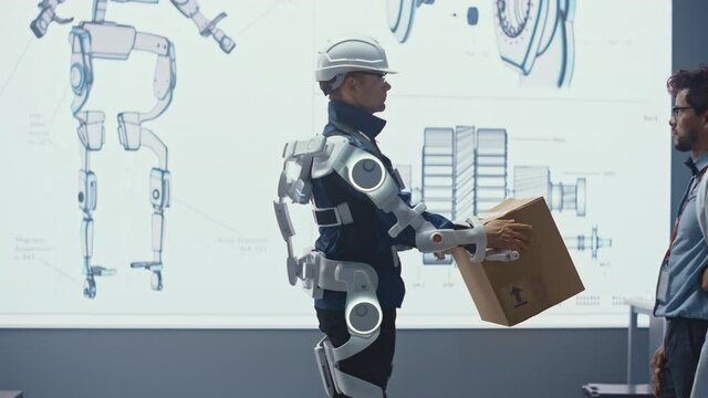 In Development Laboratory: Engineers Look at Robotics Exoskeleton Prototype Presentation with Person Testing it, Lifting Heavy Carboard Box. Designing Wearable Exosuit to Help Warehouse Workers.