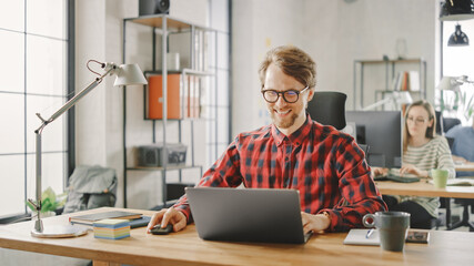 Handsome Smiling Young Man in Glasses and Shirt is Working on a Laptop in a Creative Business Agency. They Work in Loft Office. Diverse People Working in the Background. He's in Good Mood.