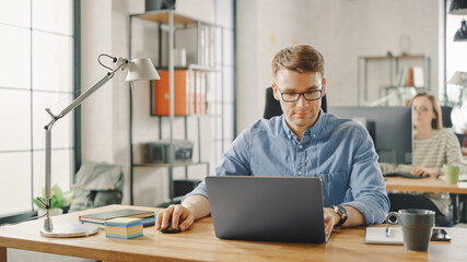 Handsome Young Man in Glasses and Shirt is Working on a Laptop in a Creative Business Agency. They Work in Loft Office. Diverse People Working in the Background. He's in Good Mood.