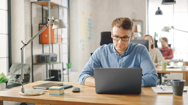 Handsome Young Man in Glasses and Shirt is Working on a Laptop in a Creative Business Agency. They Work in Loft Office. Diverse People Working in the Background. He's in Good Mood.