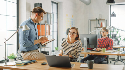 Male Colleague Sits on a Table in Front of a Young Female Cowroker while Holding a Tablet Computer with Business Notes. Friendly Atmosphere in a Creative Agency. They Discuss Business Ideas.