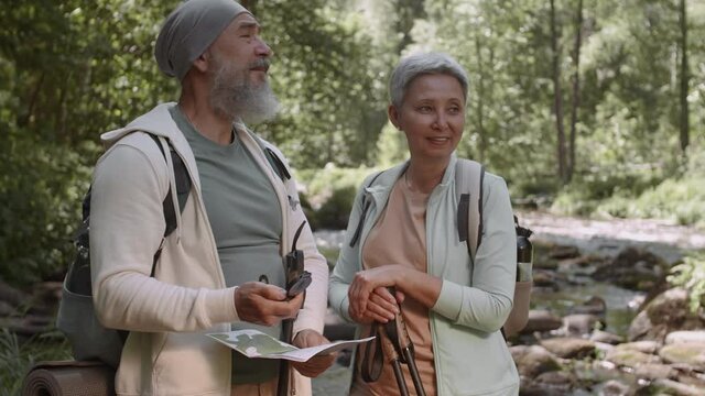 Medium shot of elderly man and woman are standing in woods near river with compass and map and talking about their route