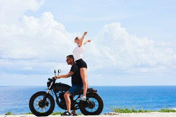 Fototapeta premium Happy young woman feeling excited while riding with boyfriend on ocean coast enjoying leisure and nature scenery, joyful couple in love satisfied with adventure during honeymoon driving motorcycle