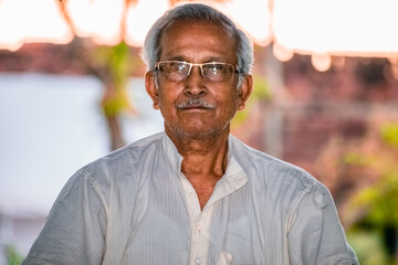 Close-up Portrait of Indian Senior Man Sitting Alone. Happy old man, wearing white dress, giving pose with style and smiling at the camera. Elderly Man Is Enjoying Retirement In Nature In Afternoon.