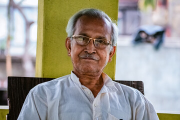 Fototapeta premium Close-up Portrait of Indian Senior Man Sitting Alone. Happy old man, wearing white dress, giving pose with style and smiling at the camera. Elderly Man Is Enjoying Retirement In Nature In Afternoon.