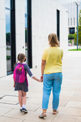 mother leads her daughter to study by the hand. walk together in the park, rear view. girl in a medical mask, in uniform, with a school bag