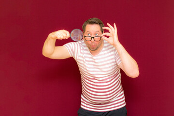 Man with eye glasses wears striped t shirt taking a magnifying glass and looking through it.Burgundy background.