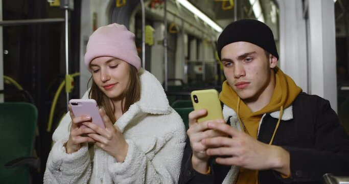 Young couple using their smartphones while sitting on seats in public transport. Good looking guy and girl phubbing while going together in tram. Concept of addiction.
