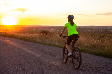 Naklejka na ściany i meble Beautiful girl cyclist riding a bike on the road towards the sunset. Nature and recreation. Hobbies and sports